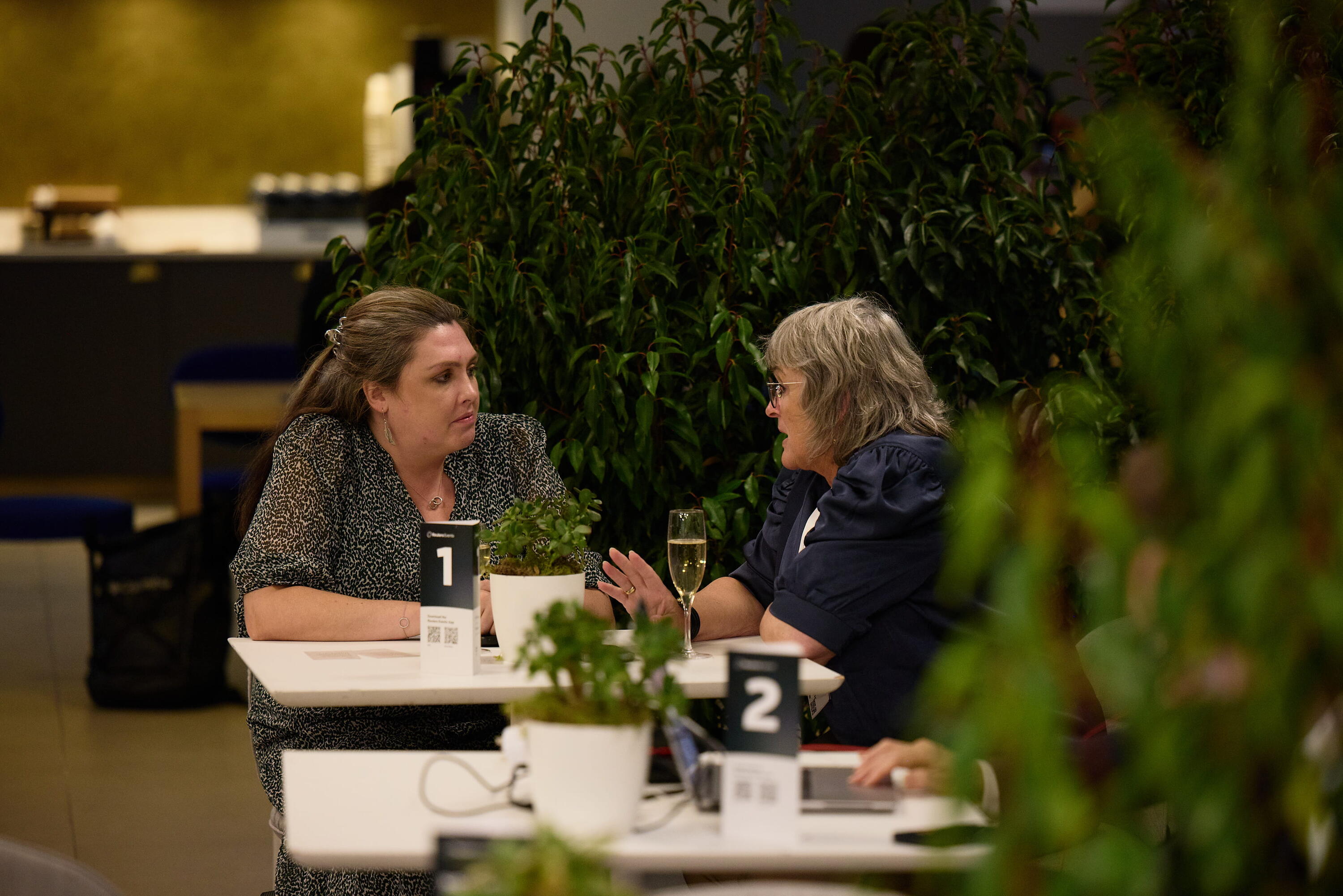 Two women engage in a one-to-one meeting at a numbered table surrounded by greenery during the Reuters Events Sustainability Europe conference.