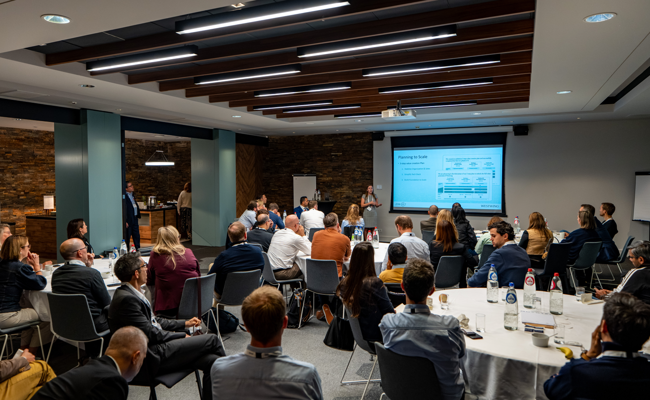 Audience seated at round tables watching a presenter speak beside a projected slide in a conference room.