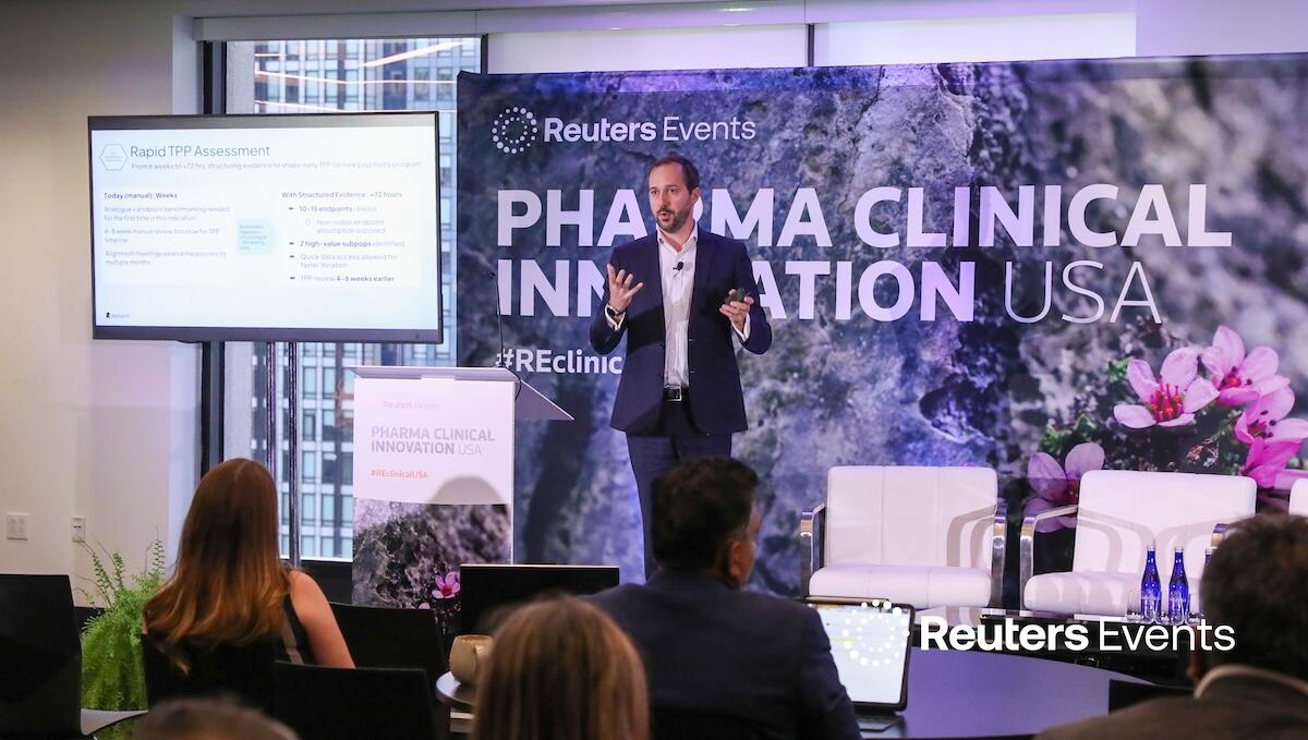 A speaker on stage at a Reuters Events conference giving a presentation to a room full of people