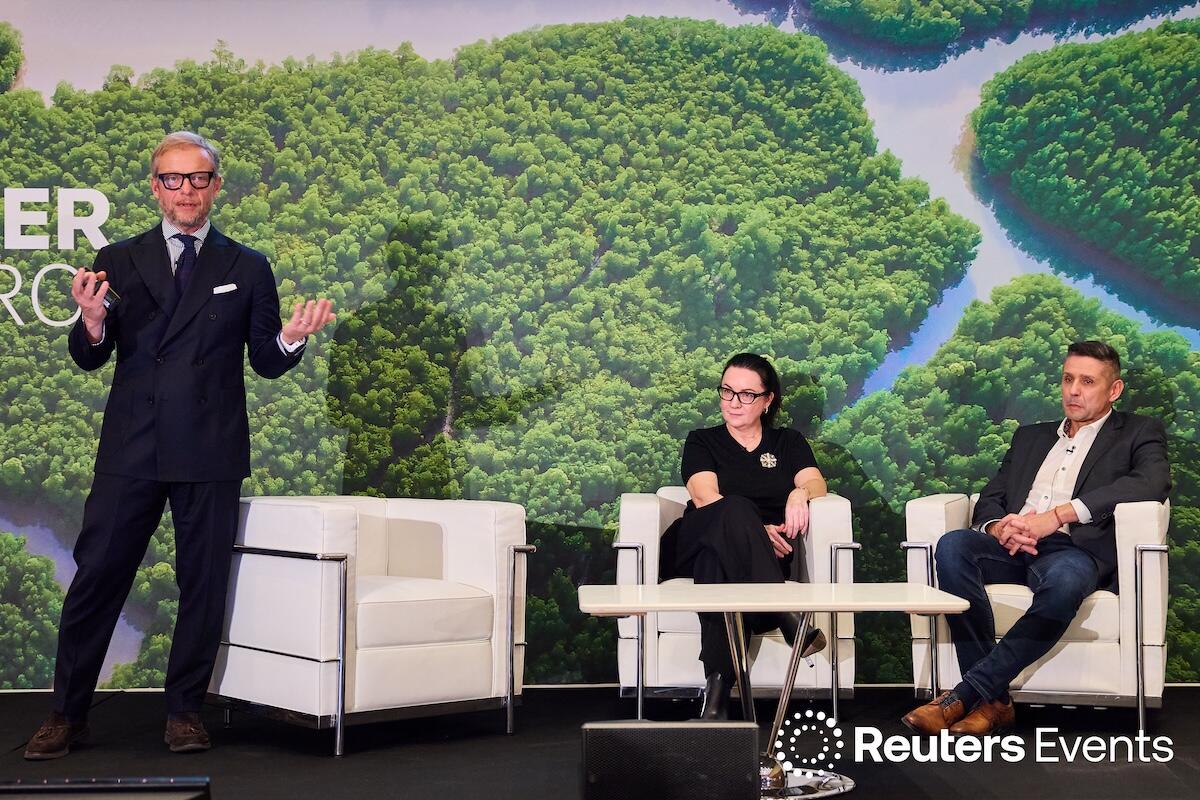 Three speakers on stage at a Reuters Events conference giving a presentation. One is standing up, hands out wide, whilst the other two are sitting in white modern chairs.