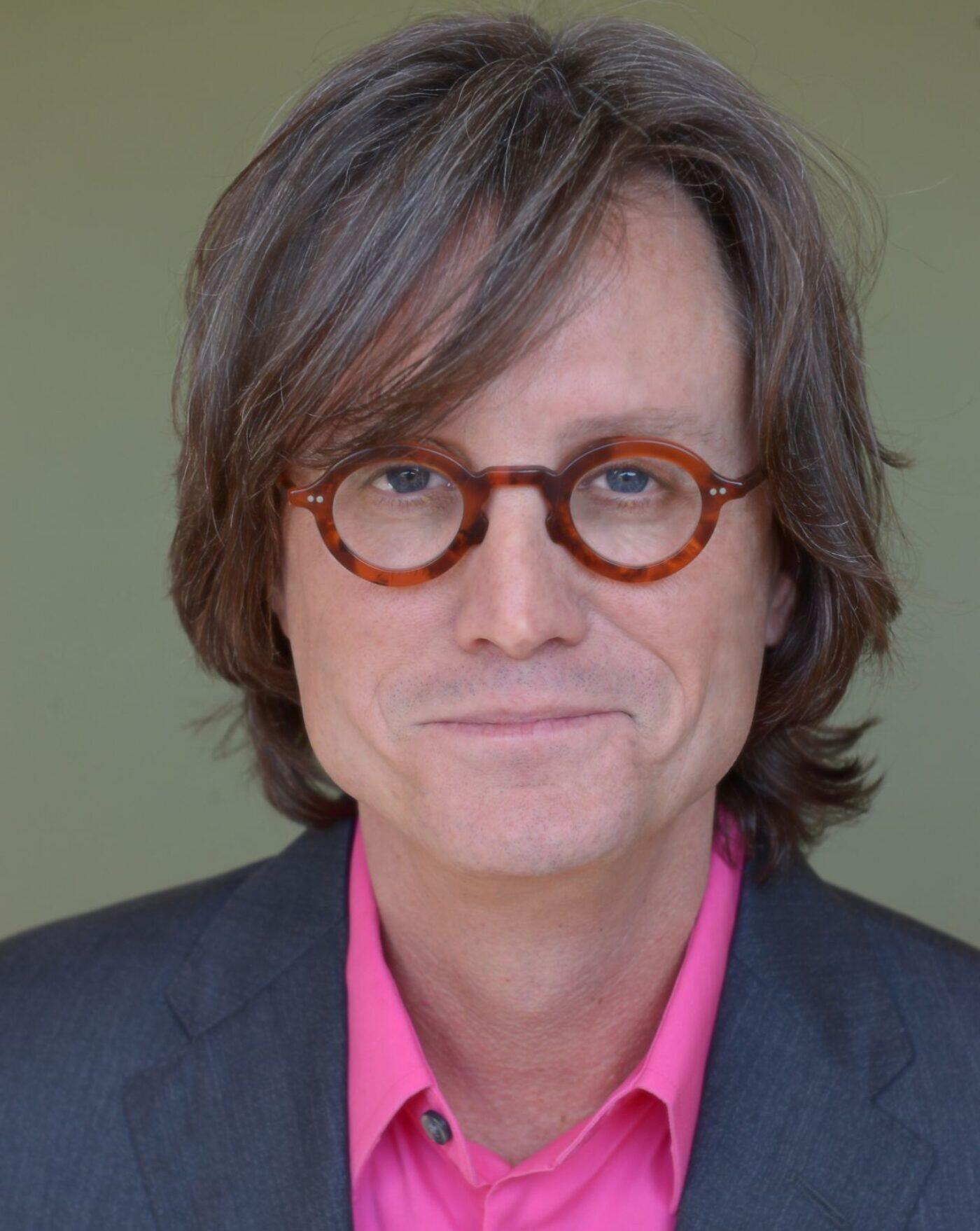 Biography portrait of Greg Cope White, a middle aged male wearing leopard print glasses and a pink collared shirt beneath a navy blue blazer