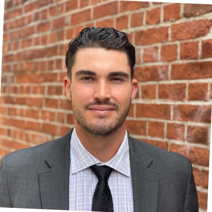 Headshot of Jordan Benty, a man wearing a checkered shirt and black tie with a gray jacket, set against a brick wall background