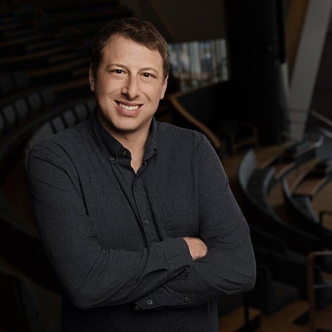 A white man with brown hair looking like a rockstar wearing a charcoal grey button down shirt. He is standing in the middle of the Ralph J Roberts Theater.    