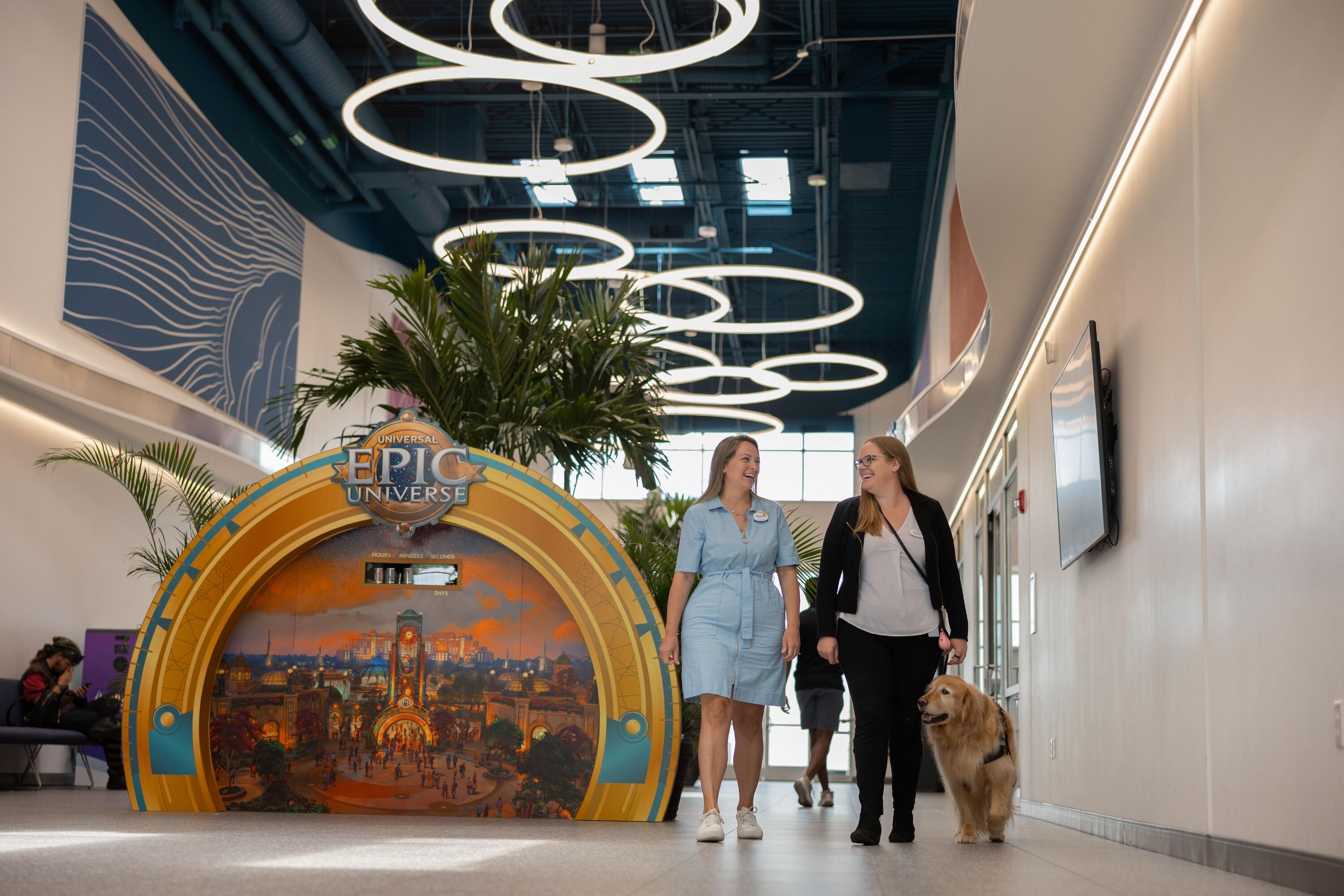 A bright, modern indoor lobby or corridor with a high ceiling featuring multiple circular ring lights suspended overhead. Two people walk side by side down the center of the hallway. One person is wearing a blue dress and white shoes and one person is wearing black pants, a white shirt, and a black sweater, accompanied by a service dog. On the left side, there is a large, colorful display with the words “Epic Universe,” showing an illustrated cityscape scene framed by a decorative archway, with tall green plants behind it. On the right wall, a mounted screen is visible along a clean, white corridor. The space has a contemporary design with soft, even lighting and a polished floor, creating an open and welcoming environment.