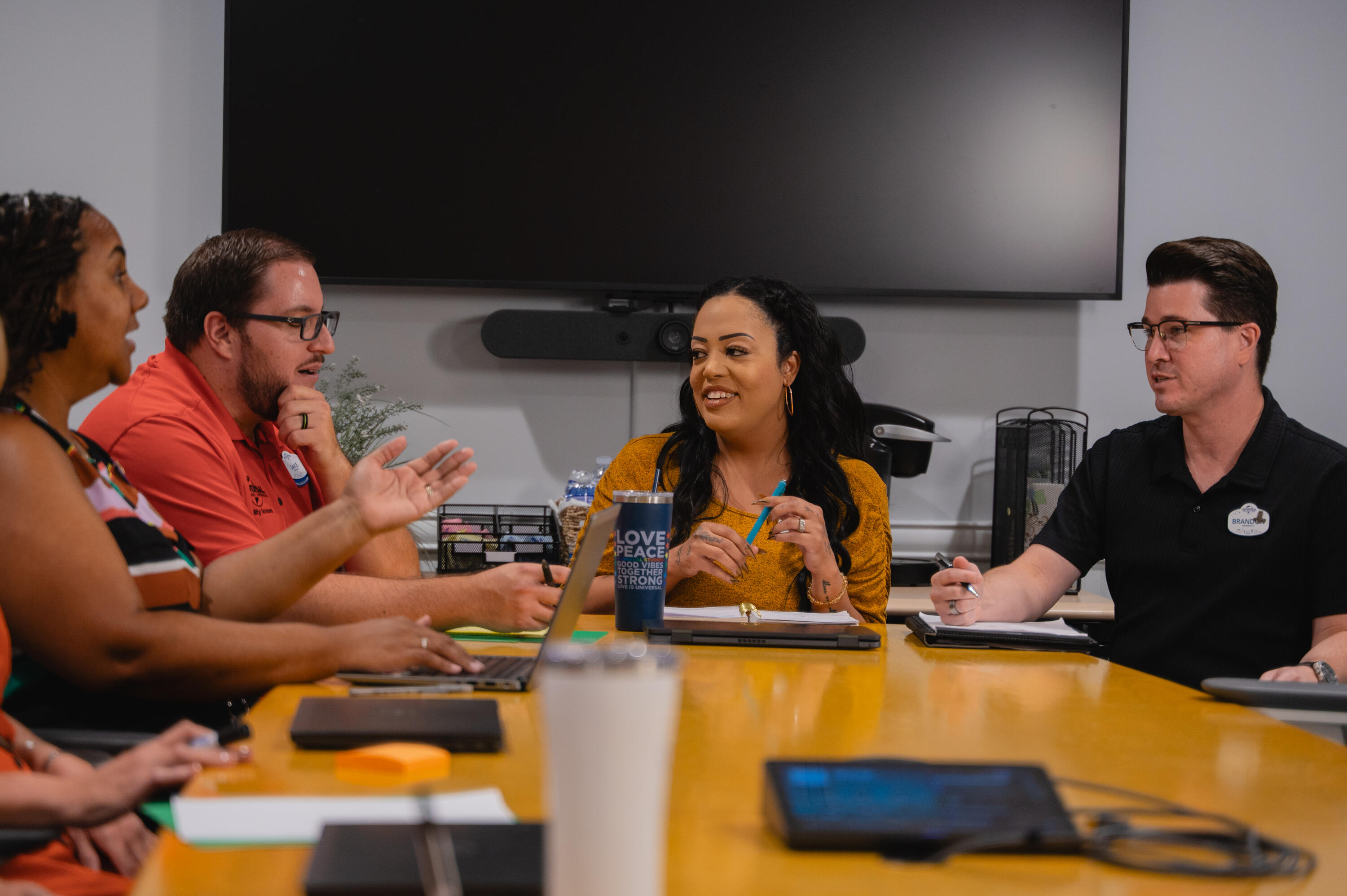 A group of five people sit around a rectangular conference table in a meeting room, engaged in discussion. Laptops, notebooks, tablets, and drinks are arranged across the glossy tabletop. A clear water bottle with printed text is placed near the center, alongside a reusable cup and other work materials. In the background, a large wall-mounted screen is turned off, with a soundbar or speaker positioned beneath it and office equipment nearby.  The lighting is soft and even, and the overall setting suggests a collaborative workplace or team meeting environment.