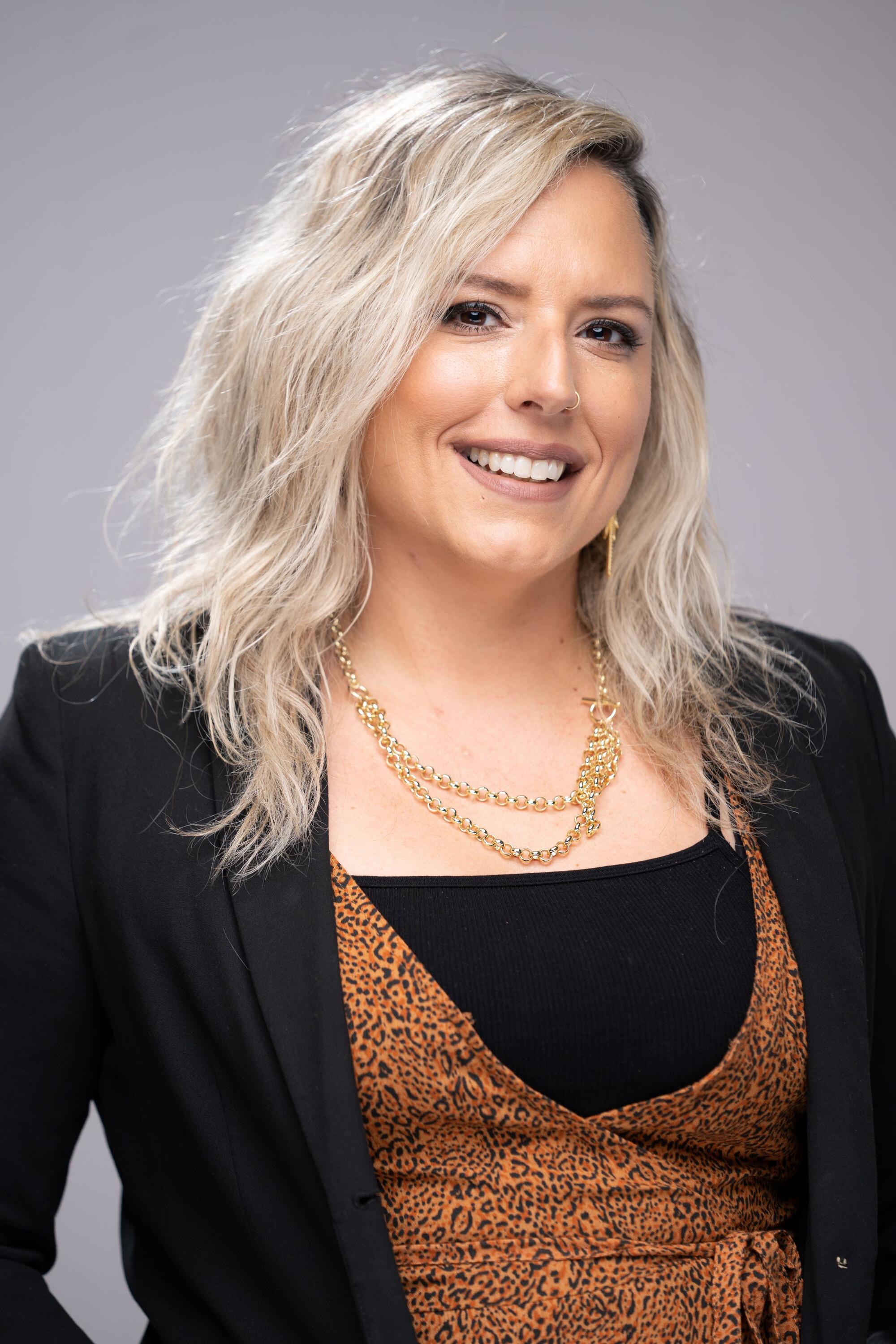 Studio headshot of a person with shoulder-length light blond hair, photographed against a light gray background. The person is wearing a black blazer over a black top and an orange-and-black patterned wrap-style dress or camisole. Gold accessories include layered chain necklaces, a long earring, and a small nose ring. The portrait is cropped from the waist up, with the person facing the camera in a professional studio setting.