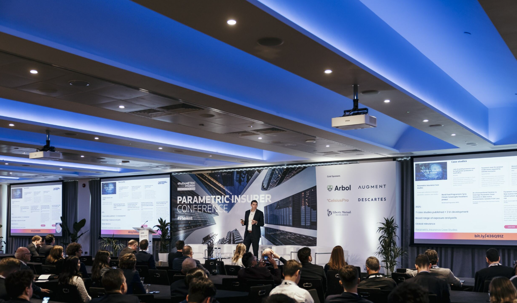 A man in a dark suit stands centre stage at the Parametric Insurer Conference, holding a small device and presenting to a large seated audience in a modern conference room with blue ambient ceiling lighting; behind him is a large branded backdrop reading "Parametric Insurer Conference #PIevent" with an aerial cityscape graphic, flanked by a gold sponsors panel displaying the logos of Arbol, Augment, CelsiusPro, Descartes, and Liberty Mutual Re; two large projection screens on either side display "Case Studies" slides with text and images; two white armchairs and a podium are visible on stage, with potted tropical plants on either side of the backdrop; an Epson projector is mounted from the ceiling above the audience, and multiple attendees are visible from behind, some holding phones or tablets.