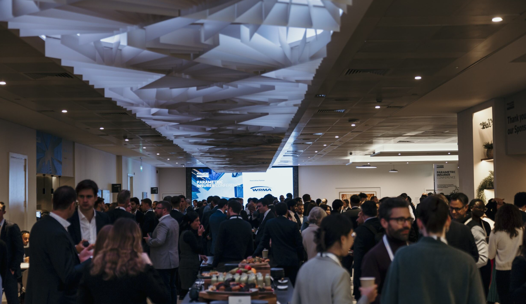 A large crowd of professionals networking at the Parametric Insurer Conference, beneath a sculptural geometric white ceiling installation, with two projection screens displaying the conference branding and WRMA logo visible in the background.