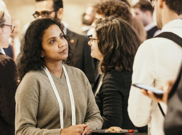 A woman in a beige V-neck sweater wearing a white conference lanyard holds a small black plate with food while engaging in conversation with a woman in a black top and glasses, surrounded by other attendees networking in a warmly lit conference venue.