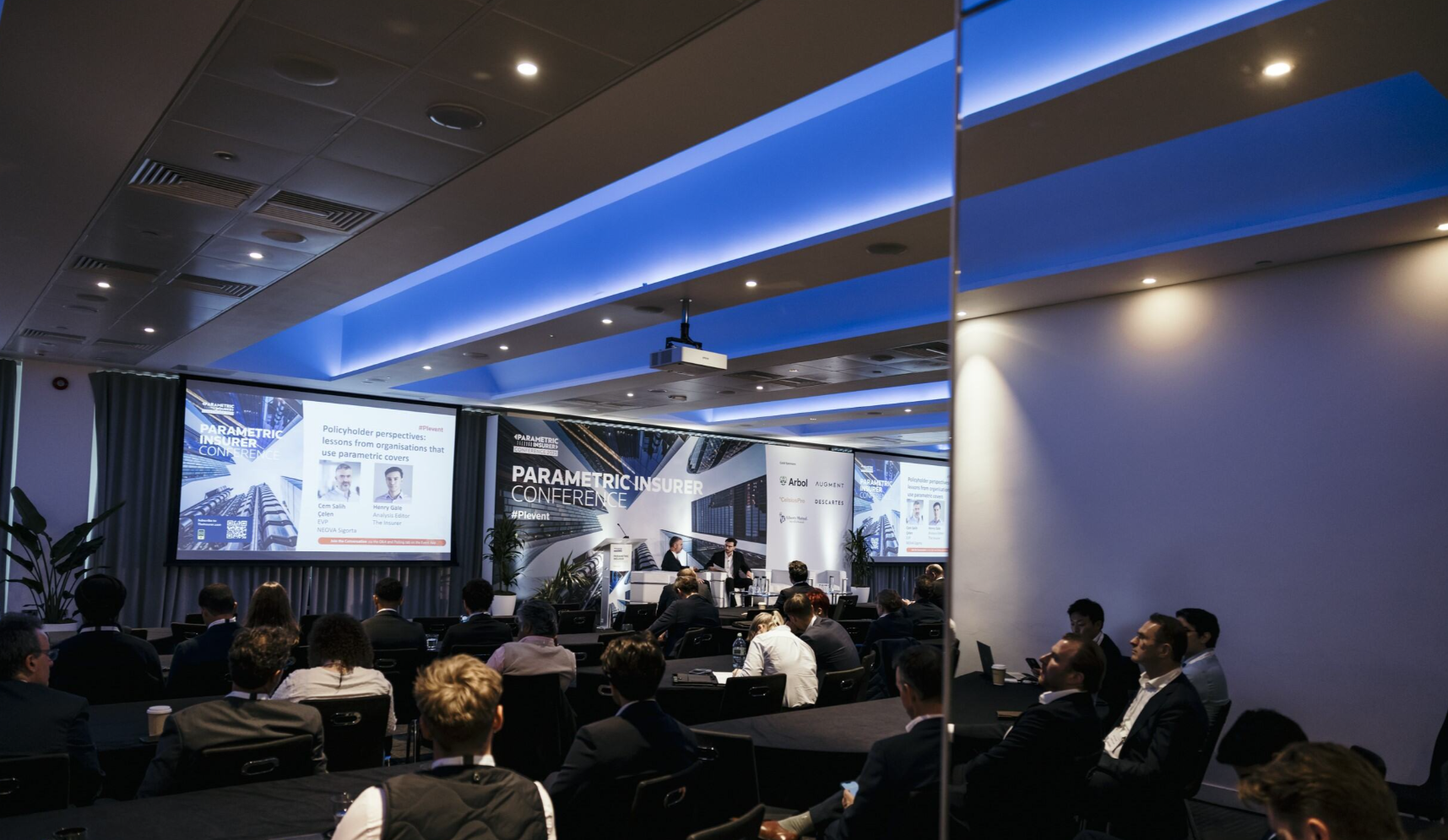 A wide-angle view of a conference room at the Parametric Insurer Conference, with a large seated audience of professionals in business attire watching two speakers on stage; the central backdrop reads "Parametric Insurer Conference #PIevent" with an aerial cityscape graphic, flanked by a gold sponsors panel displaying the logos of Arbol, Augment, CelsiusPro, and Descartes; the left projection screen displays a session slide titled "Policyholder perspectives: lessons from organisations that use parametric covers" featuring headshots and names of speakers Cem Salik, Caten, EIR, SIGMA Sigenta and Henry Gale, Analysis Editor, The Insurer; a third screen on the far right mirrors the same slide; a ceiling-mounted Epson projector is visible above the audience; the room features blue ambient ceiling lighting, dark round tables, and a potted tropical plant to the left of the stage.