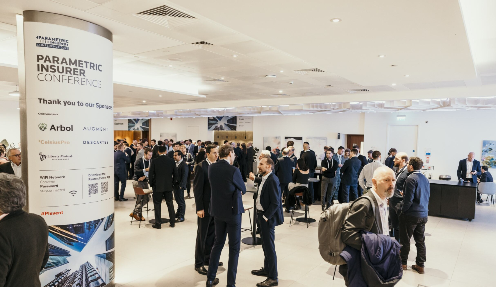 A busy networking break at the Parametric Insurer Conference 2025 in a bright, modern white-ceilinged foyer; a large branded cylindrical pillar in the foreground displays "Parametric Insurer Conference," "Thank you to our Sponsors," gold sponsor logos including Arbol, Augment, CelsiusPro, Descartes, and Liberty Mutual Reinsurance, WiFi network details (Network: Convene, Password: stayconnected), a QR code to download the Reuters Events App, and the hashtag "#PIevent" in red above an aerial cityscape graphic; dozens of professionals in dark business suits network in small groups across the open-plan space, with high round tables and bar stools scattered throughout; a bald man with a grey beard wearing a dark blazer and carrying a grey backpack and purple jacket stands in the right foreground; a dark reception or refreshments counter is visible in the far right background; two large framed photographic prints hang on the white walls in the middle background; recessed ceiling spotlights and air conditioning vents are visible overhead.