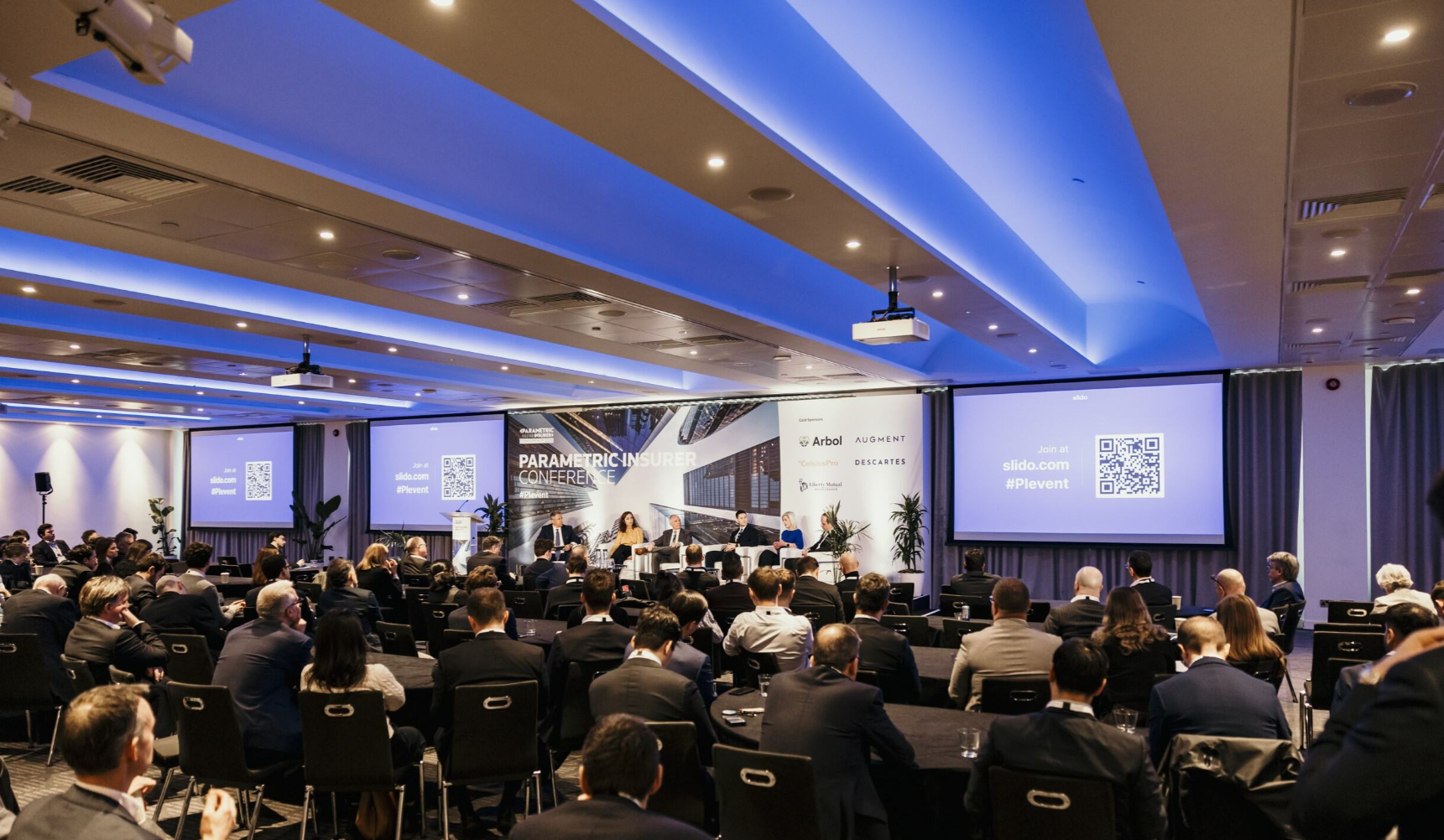 A packed conference room at the Parametric Insurer Conference, with a large audience of smartly dressed attendees seated at round tables facing a stage where five panellists are seated in white armchairs; the central backdrop displays the "Parametric Insurer Conference" branding alongside sponsor logos including Arbol, Augment, and Descartes, with three large screens showing a Slido QR code and the hashtag #PIevent, and blue ambient lighting illuminating the ceiling throughout the venue.