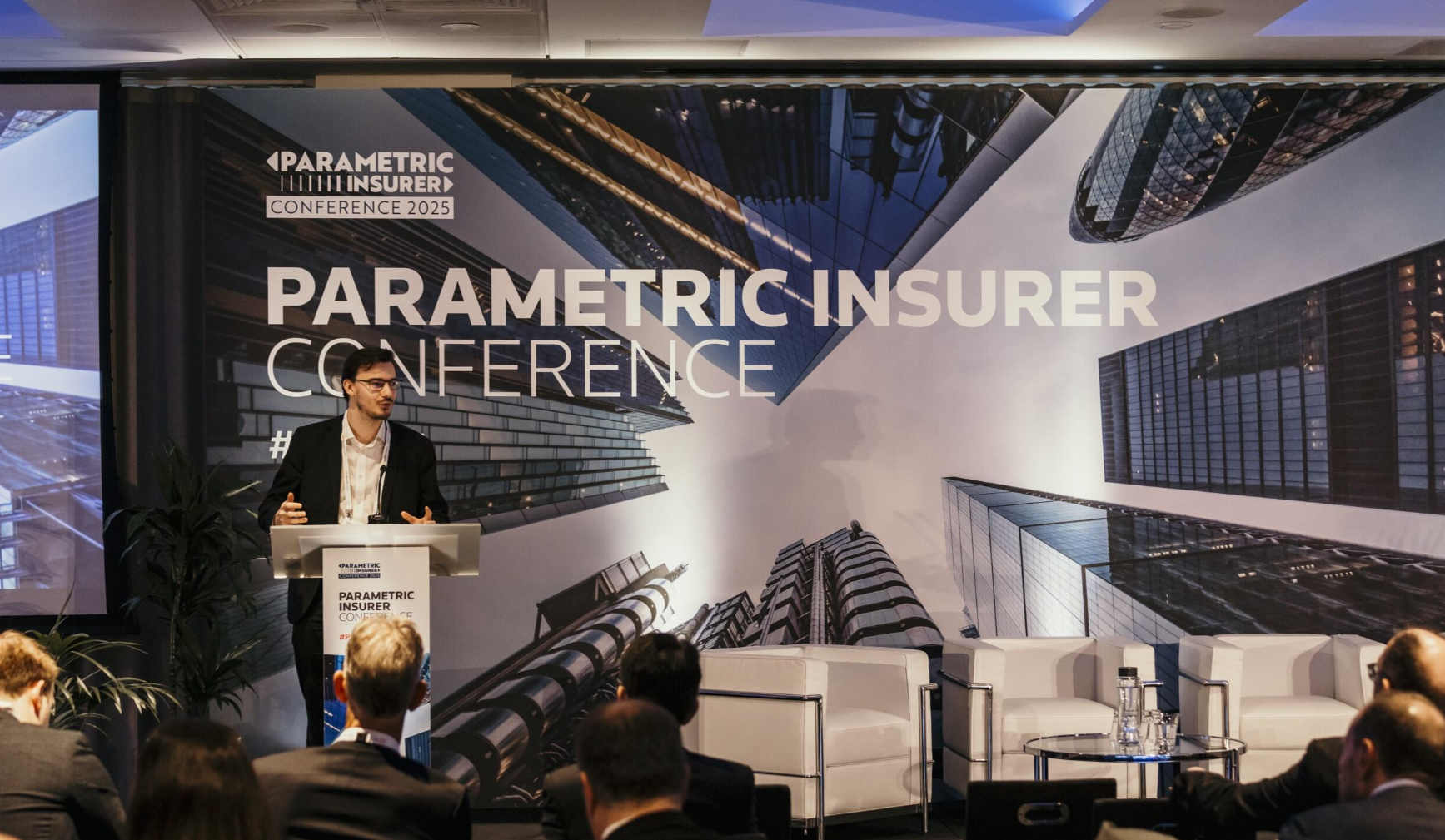 A young man in a black blazer and open-collar white shirt speaks at a glass podium at the Parametric Insurer Conference 2025, with a large branded backdrop featuring an aerial cityscape behind him, two empty white armchairs and a glass coffee table staged on the right, a projection screen on the left, and a seated audience visible in the foreground.