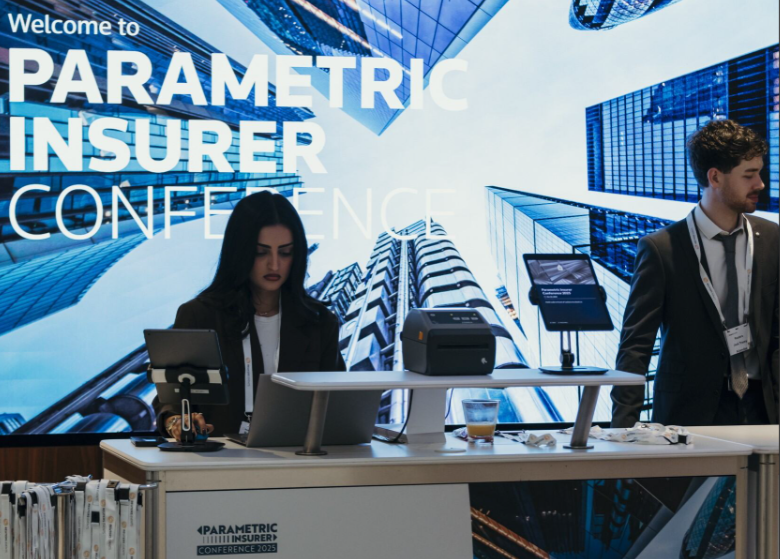 A woman with long dark hair in a black blazer and white top looks down at a laptop at the Parametric Insurer Conference 2025 registration desk, while a young man in a dark suit and tie stands to the right; a label printer, tablet, glass of juice, and lanyards are on the white desk, with a large "Welcome to Parametric Insurer Conference" branded backdrop featuring an aerial cityscape of glass skyscrapers behind them.