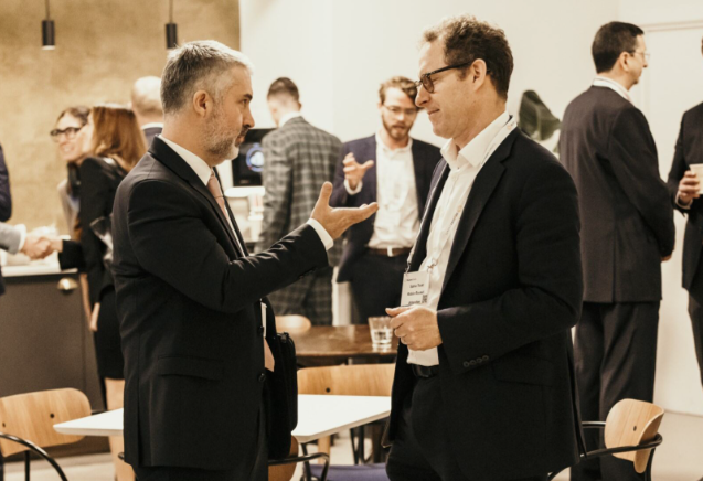 Two men in dark suits engage in conversation at a conference networking event; the man on the left gestures with his right hand while speaking, and the man on the right wearing a lanyard listens attentively, with other attendees networking and shaking hands in the softly lit background.