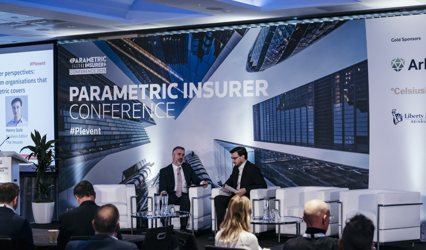 Two men in dark suits are seated in white armchairs on stage at the Parametric Insurer Conference 2025; the man on the left with a grey beard and pink tie speaks while the man on the right holds papers and listens, with a large branded backdrop displaying "Parametric Insurer Conference" and "#PIevent," a projection screen on the left showing speaker Henry Gale, Analysis Editor at The Insurer, and gold sponsor logos including Arb[ol], Celsius, and Liberty [Mutual] Re on the right; a seated audience is visible in the foreground and a potted plant flanks the left of the stage.