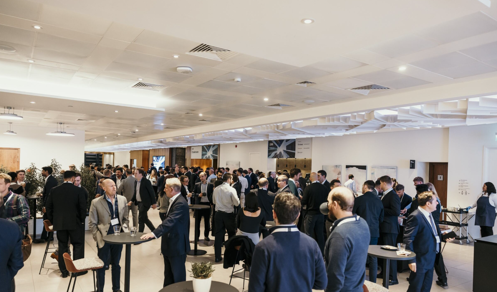 A large networking break at the Parametric Insurer Conference 2025 in a bright, modern open-plan foyer with a white tiled ceiling, recessed spotlights, and air conditioning vents; dozens of professionals predominantly in dark business suits network in small groups across the space, with round high-top black tables and brown leather bar stools scattered throughout; in the left foreground, a man in a beige blazer and open-collar shirt stands at a high-top table with two glasses of water, gesturing in conversation with a man in a dark suit; a woman in a black outfit with curly dark hair stands in the centre middle ground; a man in a navy suit with headphones around his neck and a blue lanyard stands with his back to the camera in the right centre foreground; tall green potted plants line the left wall near a wooden-panelled feature wall; two large framed black-and-white architectural photographic prints hang on the white walls in the background, one displaying the Arbol logo; a directional signage board is mounted on the far right wall near a wooden door; a small white potted olive tree sits on the floor in the foreground centre.