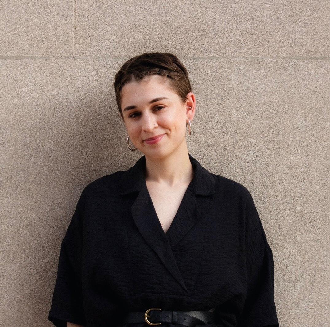 Headshot of Rebecca Fohl, a woman with short dark hair wearing a black shirt against tan cement wall