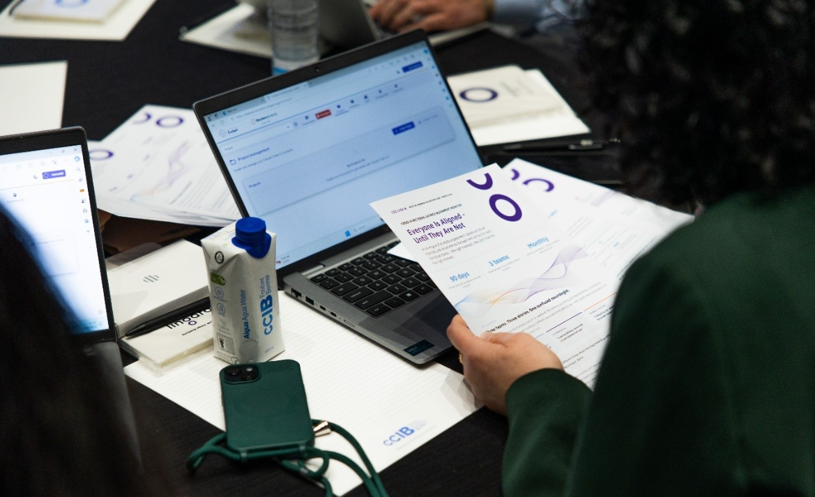 A person in a green jumper reviews CCIB-branded workshop handouts at a conference table, with a laptop, tablet, phone, and water carton nearby.