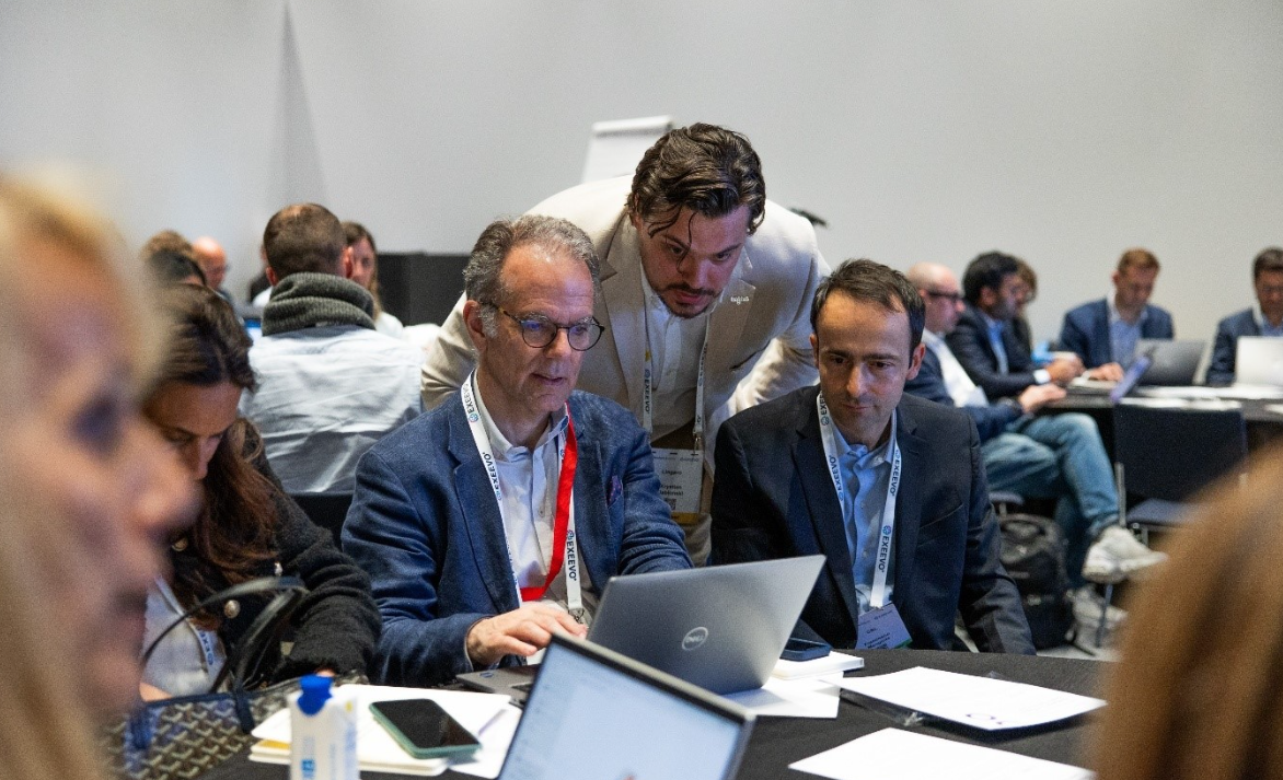 A man in a beige blazer leans over to assist two seated male attendees reviewing content on a Dell laptop at a conference workshop. The seated men wear lanyards and blazers. The room is filled with other attendees working at tables, with laptops, papers, and phones visible. A flipchart stand is in the background.