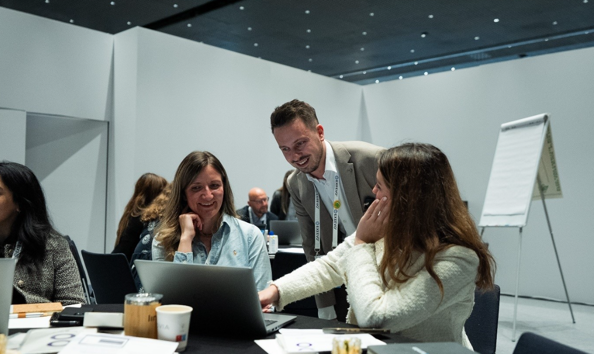 A man in a beige blazer leans over to assist two seated attendees reviewing content on a Dell laptop at a conference workshop. The seated men wear lanyards and blazers. The room is filled with other attendees working at tables, with laptops, papers, and phones visible. A flipchart stand is in the background.