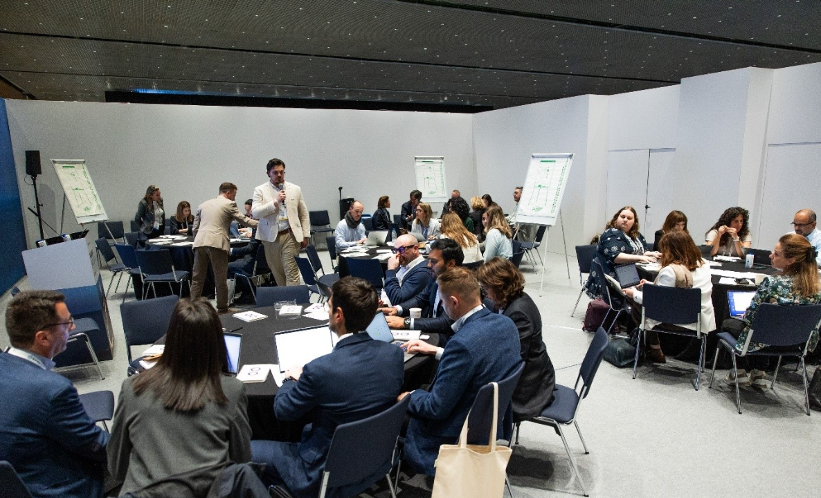 A large conference workshop session with approximately 30 attendees seated at round tables in groups, working on laptops and printed materials. A man in a beige blazer stands at the front holding a microphone, facilitating the session. Three flipchart stands displaying green-printed diagrams are positioned around the room. Attendees are dressed in business attire, with laptops, papers, and drinks on the tables. A tote bag hangs on a chair in the foreground. The room has a dark ceiling with subtle lighting and white walls.