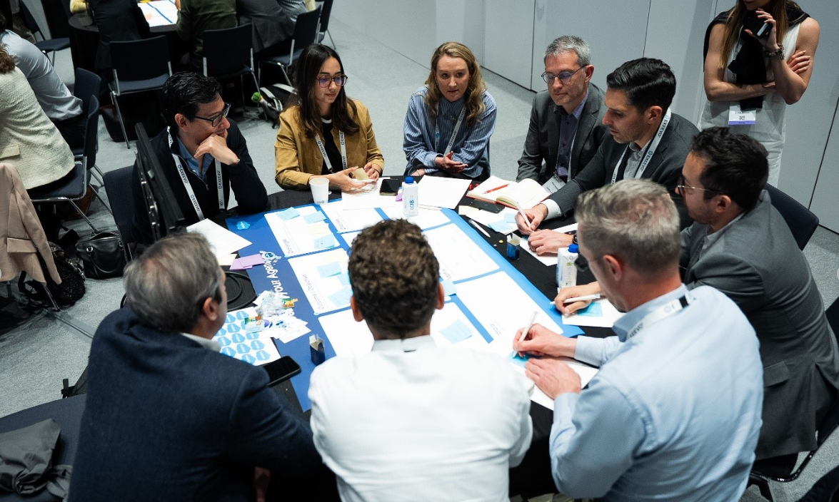 A group of approximately nine conference attendees seated around a round table covered with a blue tablecloth, engaged in an active group discussion. The table is covered with printed worksheets, sticky notes, pens, a water bottle, and a plastic cup. A woman in a blue striped shirt with a lanyard is speaking, while others listen and take notes. Attendees wear business attire including a mustard yellow blazer, grey suits, a navy blazer, and a light blue shirt. All visible attendees wear conference lanyards. Other groups are visible working at tables in the background.