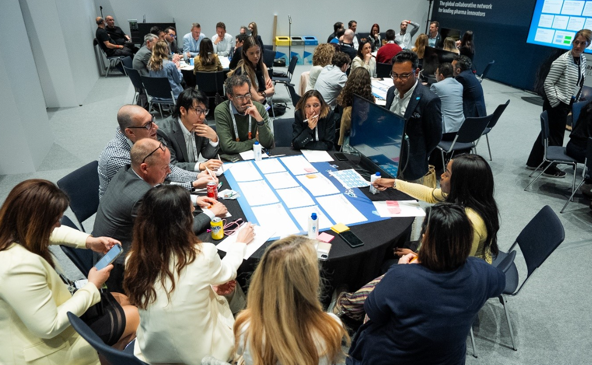 A wide-angle overhead view of a busy conference workshop session. In the foreground, approximately ten attendees are seated around a large round table covered with a blue grid worksheet, sticky notes, printed papers, a water bottle, a can of Coca-Cola, and a yellow NYC mug. Attendees are engaged in active discussion, wearing business casual attire including a green jumper with a lanyard, a grey suit, a white blazer, a cream blazer, and a navy outfit. A man in a dark suit stands beside the table facilitating. In the background, multiple other groups are seated at round tables engaged in similar workshop activities. A blue branded display screen on the right reads "The global collaborative network for leading pharma innovators." A woman in a black and white striped blazer stands near the screen. The room has white walls, grey flooring, and dark ceiling lighting rigs.