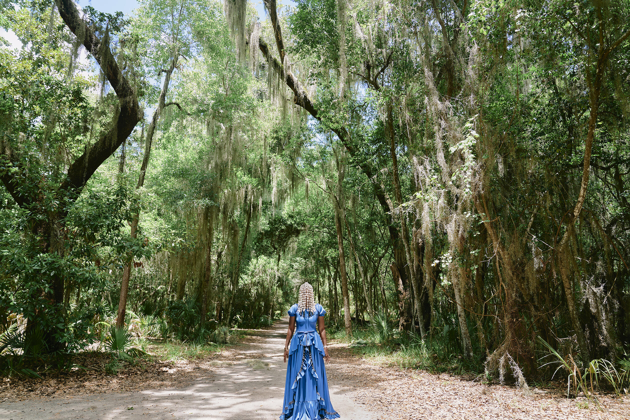 A Black woman in a long and flowing, blue dress stands on a dirt pathway surrounded by a forest of tall, mossy trees. Her beaded hair drapes over her eyes, concealing most of her face. 